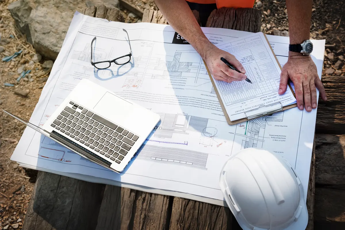 Plans de chantier, ordinateur portable et feuille de suivi posés sur une table de chantier